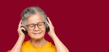 Kırmızı arka planda dururken gülümseyerek en sevdiği şarkıyı dinlemek için kısa gri saçlı, kablosuz kulaklık takan neşeli, yaşlı Asyalı kadın. Yaşlılar ve rahatlama konsepti