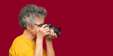 Kırmızı arka planda dikilirken dijital kamerayla fotoğraf çeken mutlu yaşlı kadın. Yaşlanmış insanlar fotoğrafçı ve rahatlama konsepti