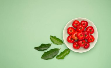 Beyaz bir kabın içinde olgun kırmızı acerola kirazları ve yeşil arka plana saçılmış yeşil yapraklar. Yüksek C vitamini ve antioksidan meyveler. Mesaj için yer var. Yakın plan. Sağlıklı meyve kavramı