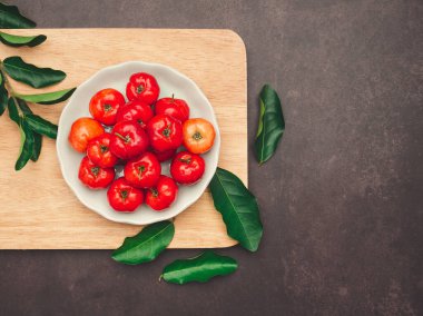 Beyaz bir tabakta olgun kırmızı acerola kirazları ve tahta kesme tahtasına saçılmış yeşil yapraklar. Yüksek C vitamini ve antioksidan meyveler. Mesaj için yer var. Üst Manzara. Yakın plan. Sağlıklı meyve kavramı