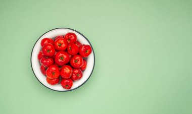 Yeşil zemin üzerinde beyaz bir tabakta olgun kırmızı acerola kirazlarının üst görüntüsü. Yüksek C vitamini ve antioksidan meyveler. Mesaj için yer var. Yakın plan fotoğraf. Sağlıklı meyve kavramı