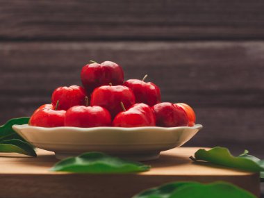 Olgun kırmızı acerola kirazları bir tabakta ve yeşil yapraklar tahta kesme tahtasına saçılmış. Yüksek C vitamini ve antioksidan meyveler. Mesaj için yer var. Yakın plan. Sağlıklı meyve kavramı