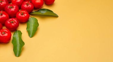 Sarı arka planda olgun kırmızı acerola kirazları ve yeşil yaprakların üst görüntüsü. Yüksek C vitamini ve antioksidan meyveler. Mesaj için yer var. Yakın plan fotoğraf. Sağlıklı meyve kavramı