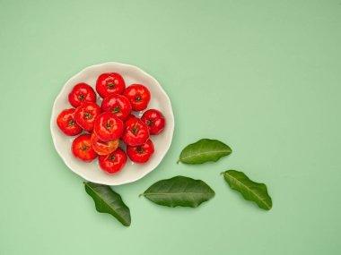 Yeşil zemin üzerinde yeşil yapraklar ve beyaz bir tabakta olgun kırmızı acerola kirazlarının üst görüntüsü. Yüksek C vitamini ve antioksidan meyveler. Mesaj için yer var. Yakın plan fotoğraf. Sağlıklı meyve kavramı
