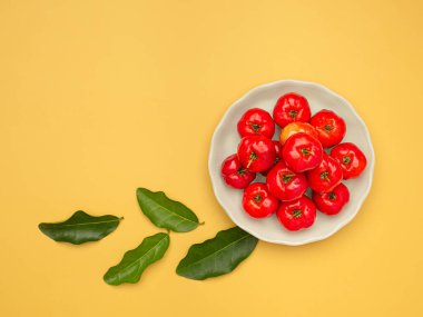 Beyaz bir tabakta olgun kırmızı acerola kirazları ve turuncu arka planda yeşil yapraklar. Yüksek C vitamini ve antioksidan meyveler. Mesaj için yer var. Yakın plan fotoğraf. Sağlıklı meyve kavramı
