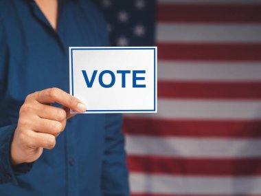 Oy kullanma hakkını kullanma kampanyası. Amerikan bayrağı arka planında dururken elinde VOTE yazılı bir kağıt tutan seçmenin yakın çekimi. Özgürlük demokrasi kavramı. Yakın plan fotoğraf.