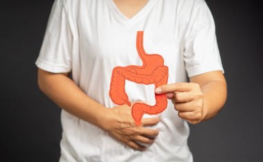 Kolon sembolü tutan ve karın ağrısına dokunan elin yakın plan resmi. Gri arka planda dururken karın ağrısı çekiyor.