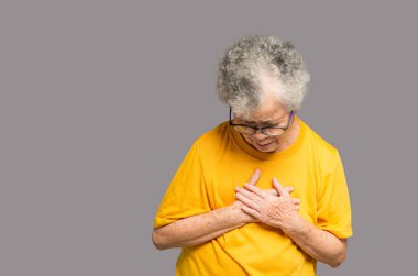 Turuncu günlük giyinmiş yaşlı bir kadın stüdyoda gri arka planda dururken sol göğsünde ağrı hisseder. Yakın plan fotoğraf. Mesaj için yer var. Yaşlılar, sağlık ve sağlık hizmetleri kavramı