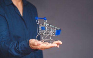 Mavi gömlekli bir adam, gri arka planda dururken avucunda küçük bir alışveriş arabası var. Çevrimiçi alışveriş kavramı. Yakın plan fotoğraf.