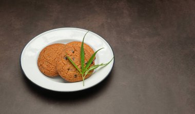 Esrar parçalı çikolatalı kurabiyeler, beyaz bir tabakta marihuana yaprağı ve klasik bir arka plan. Kenevir bitkisiyle yemek pişirme kavramı