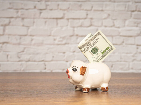 Концепция сберегательных денег. Прекрасная копилка с банкнотой в долларах США на деревянном столе на фоне стены из белого кирпича. Пространство для текста
