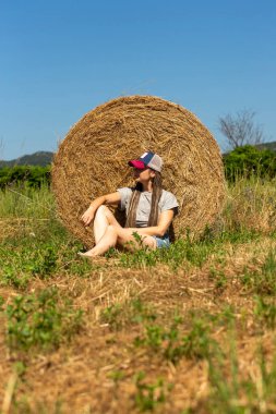 Genç bir kadın samanlıkta saman balyasında oturur, güneş ışığında güneşlenir, arka planda dağlar, şapkalı kadın saman balyasında oturur, saman yığınının üzerinde uzanan saman şapkalı doğal sahne kadını