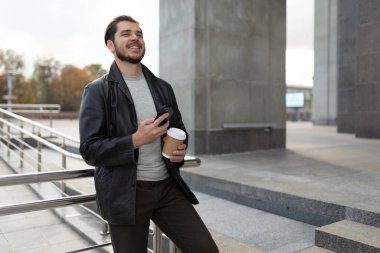 Elinde bir fincan kahveyle bir şehir binasının yanında yüzünde geniş bir gülümseme olan bir cep telefonuyla genç bir adam..