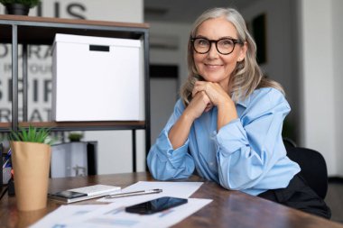 Elli yaşındaki kadın kafası başarılı bir şekilde masaya oturur ve elleri kilitlenmiş bir şekilde kameraya bakar..
