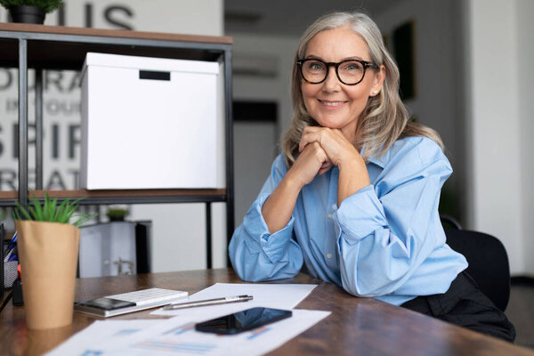 Успешно пятидесятилетняя женщина садится за стол и смотрит в камеру с руками, зажатыми в замок.