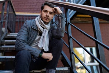 Sonbahar kıyafetleri içinde göğsünde bir eşarp olan genç bir adam ofisin girişindeki metal merdivenleri düşünerek oturur..