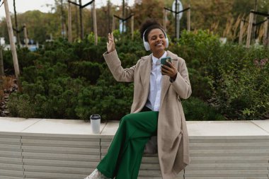 Afro-Amerikan kulaklıklı kız öğrenci elinde telefonla şehir parkının yanında oturmuş müzik dinliyor..