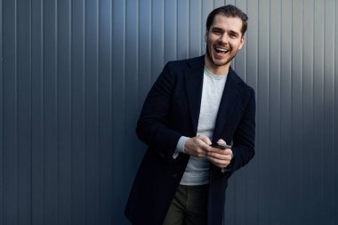 Geniş gülüşlü beyaz bir adam gri bir duvarın arkasında elinde bir telefon tutuyor..