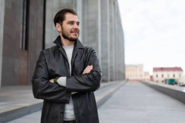 Elleri göğsünde beyaz bir adam gri bir binanın arka planında duruyor..