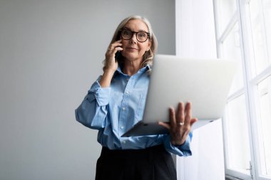 Dizüstü bilgisayarı olan başarılı bir kadın yönetici ofis penceresinde cep telefonuyla konuşuyor..