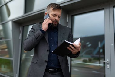 Başarılı iş adamı ofisin girişinde elinde not defteriyle cep telefonuyla konuşuyor..