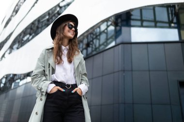 Güneş gözlüklü, siyah şapkalı, modern bir binanın arka planında genç, karizmatik bir kız..