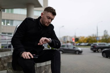 Siyah ceketli güçlü bir adam dışarıda bir toplantı bekliyor, saatine bakıyor..