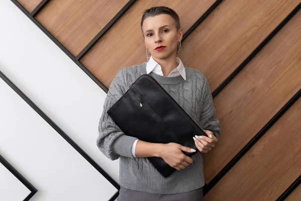 A business woman with a short haircut stands with a folder for documents on the background of a wall in the office.