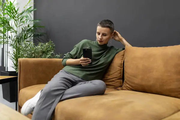 an adult woman with a short haircut sits at home on the couch with a mobile phone in her hands.