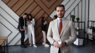 Şirketin başkanı, ön planda modern bir ofiste ciddi bir yüz ifadesiyle duruyor ve kameraya iş adamlarıyla iletişim kuran kişilerin arka planına bakıyor..