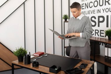 Ofiste kısa saçlı, elinde belgelerle bir kadın noter masanın yanında duruyor..