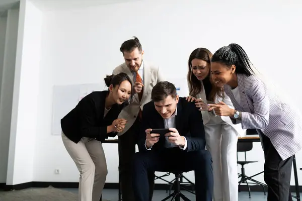 a team of professional office workers stands around a colleague playing on a mobile phone in the office.
