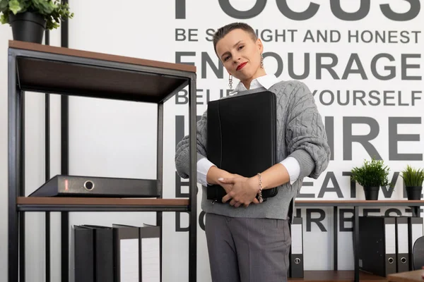 a middle-aged businesswoman with a short haircut stands a leather folder for documents in her hands against the backdrop of the office.
