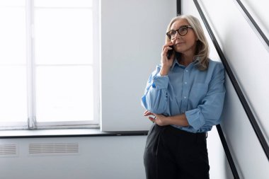 Mavili beyaz iş kadını ofisteki cep telefonunda duvara yaslanmış bir şekilde konuşuyor..