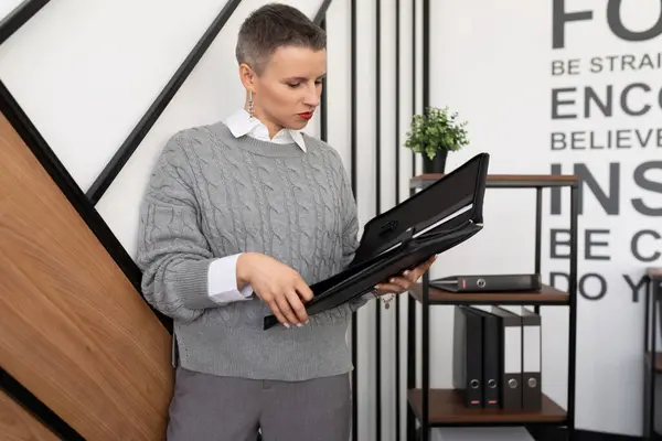 mature caucasian businesswoman with short haircut holding leather folder for documents in office.