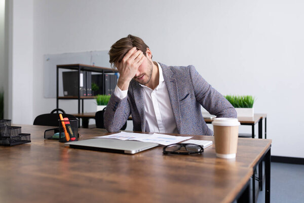 tired upset businessman sits in front of the desktop in the office covering his face with his palm.