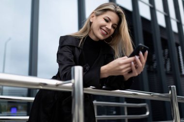 Cam bir cephenin arka planında elinde telefonla gülümseyen şık bir iş kadını..