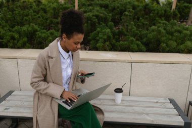 Bilgisayarda çalışan Afro-Amerikalı genç bir kadın akıllı telefon ekranına bakarak ofisin yanındaki parkta oturuyor..