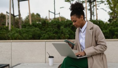 Yetişkin Afro-Amerikan iş kadını dışarıda dizüstü bilgisayarda çalışıyor. Elinde telefon var..