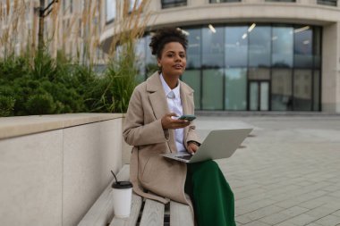 Yetişkin iş kadını Afro-Amerikan kadın ofis merkezinin yanında dizüstü bilgisayarı ve elinde bir telefonla oturuyor..