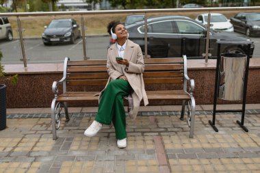 Yüzünde geniş bir gülümseme olan yetişkin Afro-Amerikalı kadın dışarıda otururken kulaklıkla müzik dinliyor..
