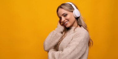 Sarı saçlı, beyaz kulaklıklı genç bir kadın turuncu arka planda müzik dinliyor..