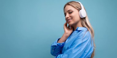 Kablosuz büyük kulaklıklı çekici bir kızın yatay fotoğrafı mavi arka plandaki bir müzik listesinden gelen müziği zevkle dinliyor..