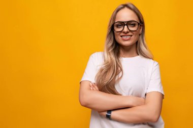 Gözlüklü ve beyaz tişörtlü kendine güvenen kız kollarını sarı arka planda boş bir alanda kavuşturdu..