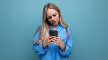 Büyülenmiş sarışın kadın mavi parlak bir arka planda elinde bir telefon tutuyor..