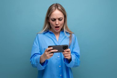 Şaşkın sarışın kız elinde akıllı telefonu, izole edilmiş mavi arka planda tutarken şok oldu..