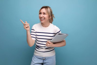 Çizgili kazaklı çekici sarışın bir kadının elinde mavi bir arka planda dizüstü bilgisayar tutarken yakın plan fotoğrafı..