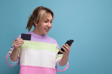 Şık, sarışın, günlük kıyafetlerle gezen, elinde kredi kartı olan, mavi arka planda fotokopi alanı olan, akıllı telefonlu bir kadın..