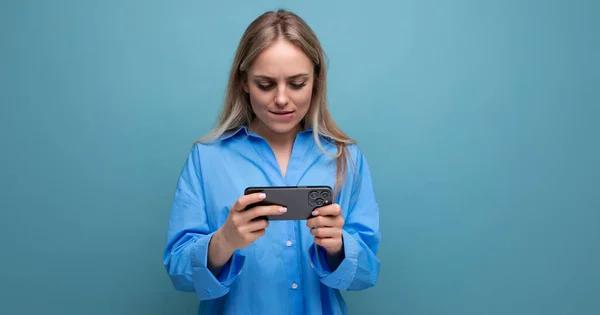 horizontal photo of a lucky charming blonde girl holding a smartphone in her hands on an isolated blue background.
