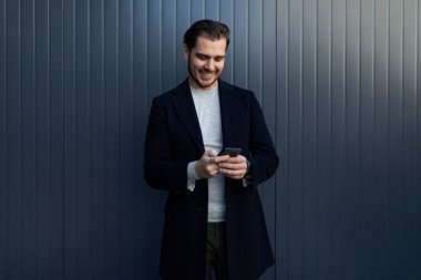 Bir binanın arka planında akıllı telefonuyla gülümseyen Avrupalı bir adamın portresi.